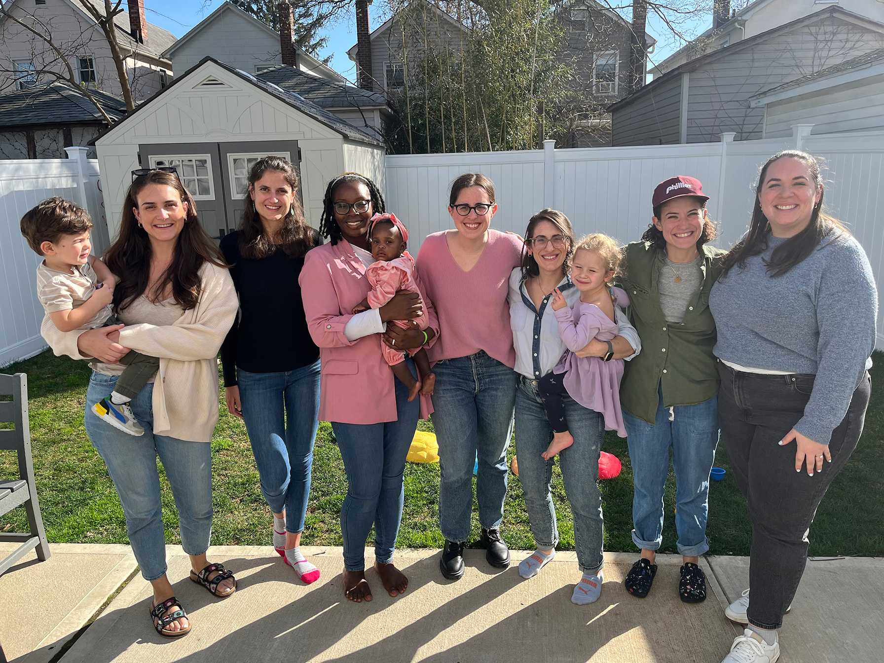Friends and their children smiling in a sunny backyard with a white fence and small shed behind them.