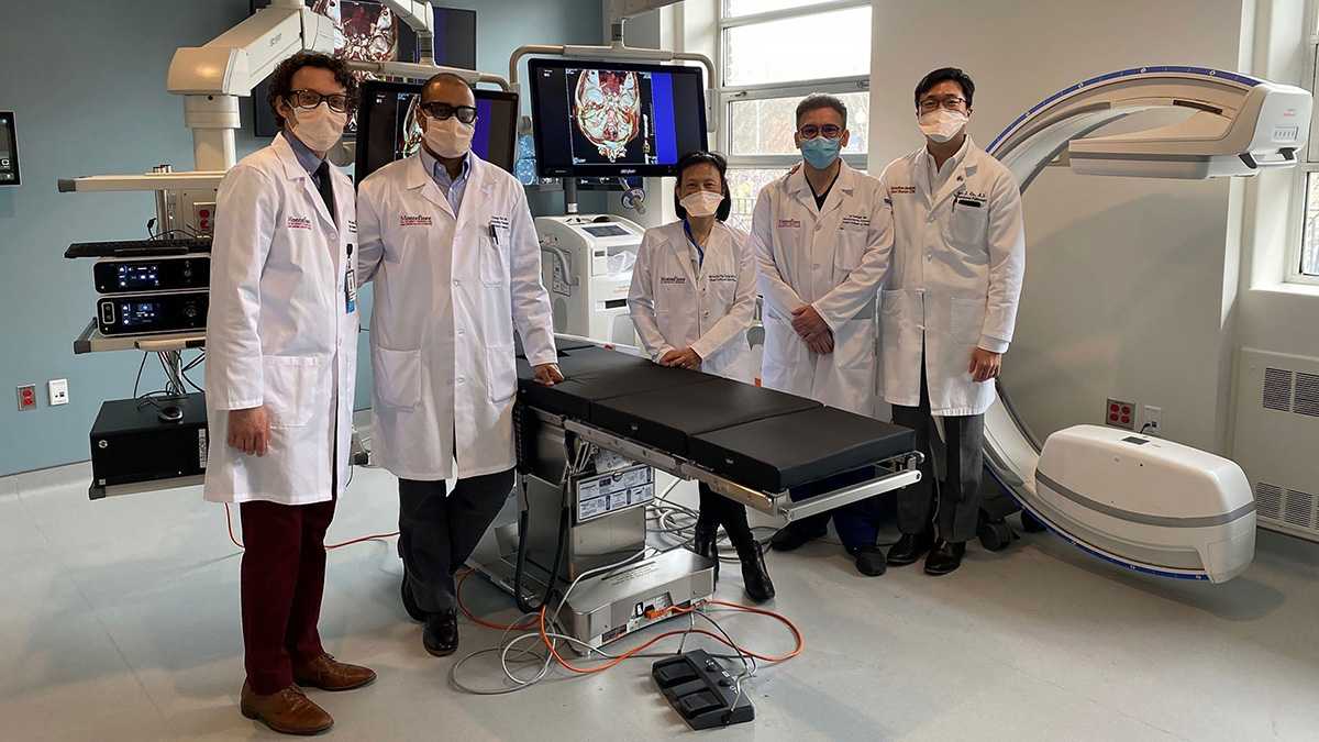 Five doctors in lab coats and face masks standing in a bronchoscopy suite with imaging monitors and surgical equipment.