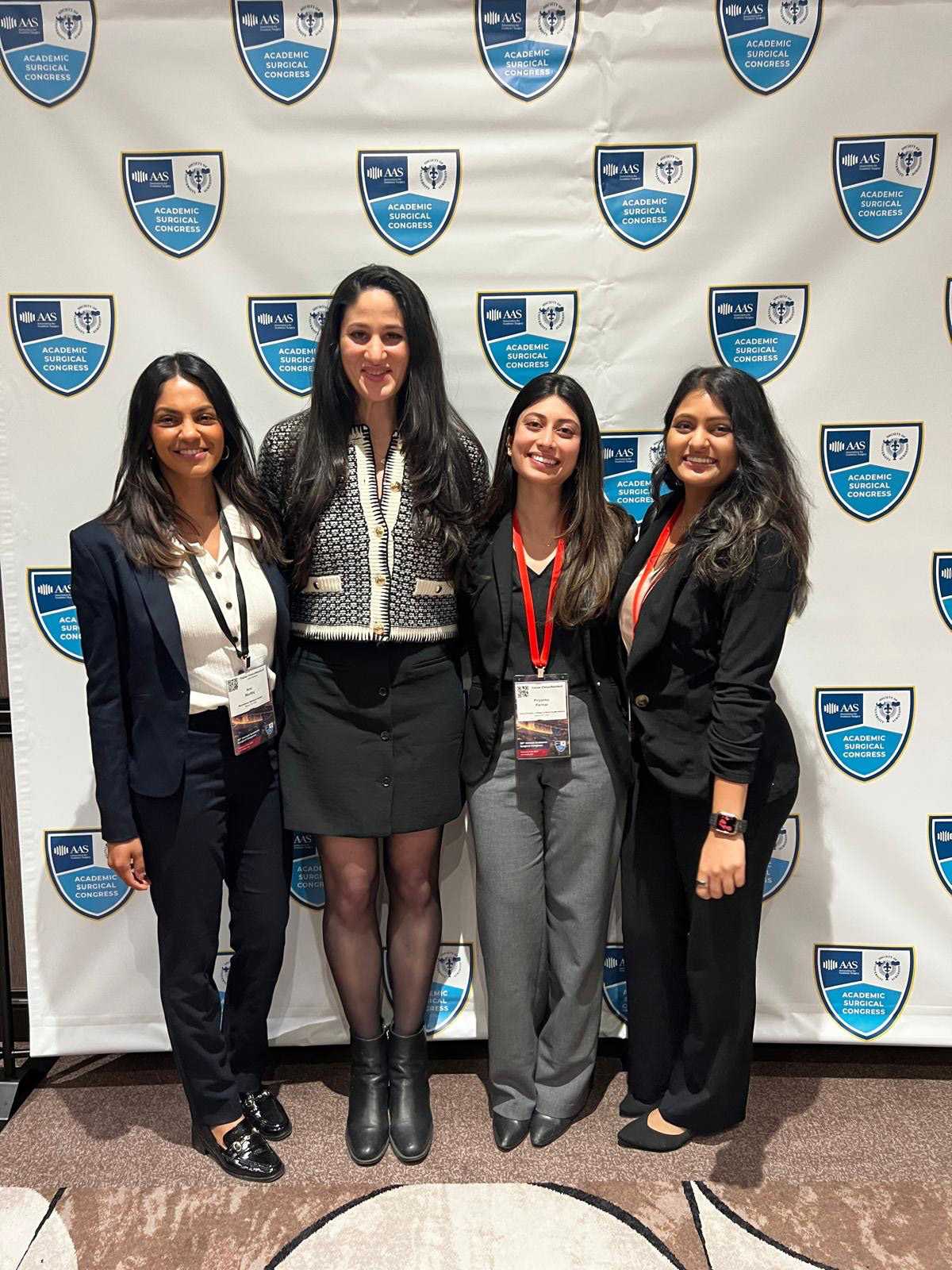 Four women dressed in business attire posing in front of an Academic Surgical Congress step-and-repeat backdrop.