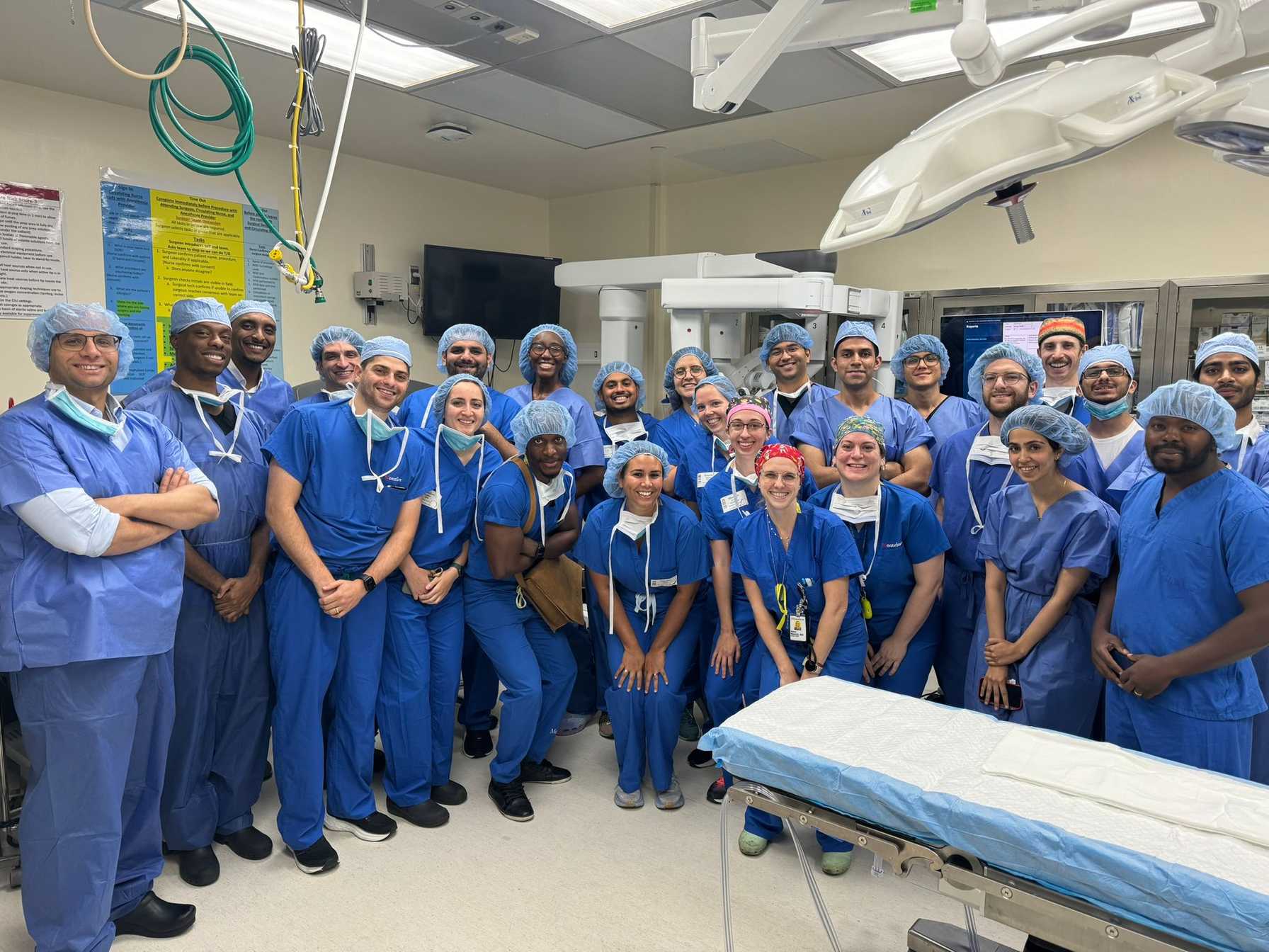 Large group of medical professionals in blue surgical scrubs and caps posing together in an operating room.