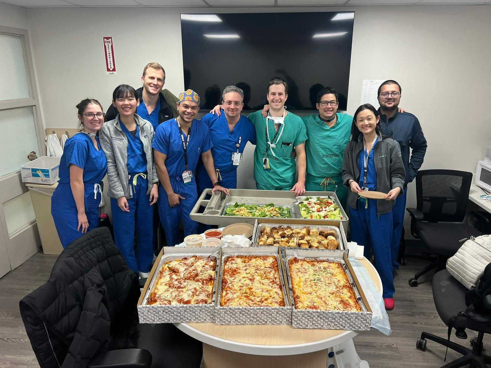 Medical team in scrubs gathered around a table filled with trays of food in a hospital break room.