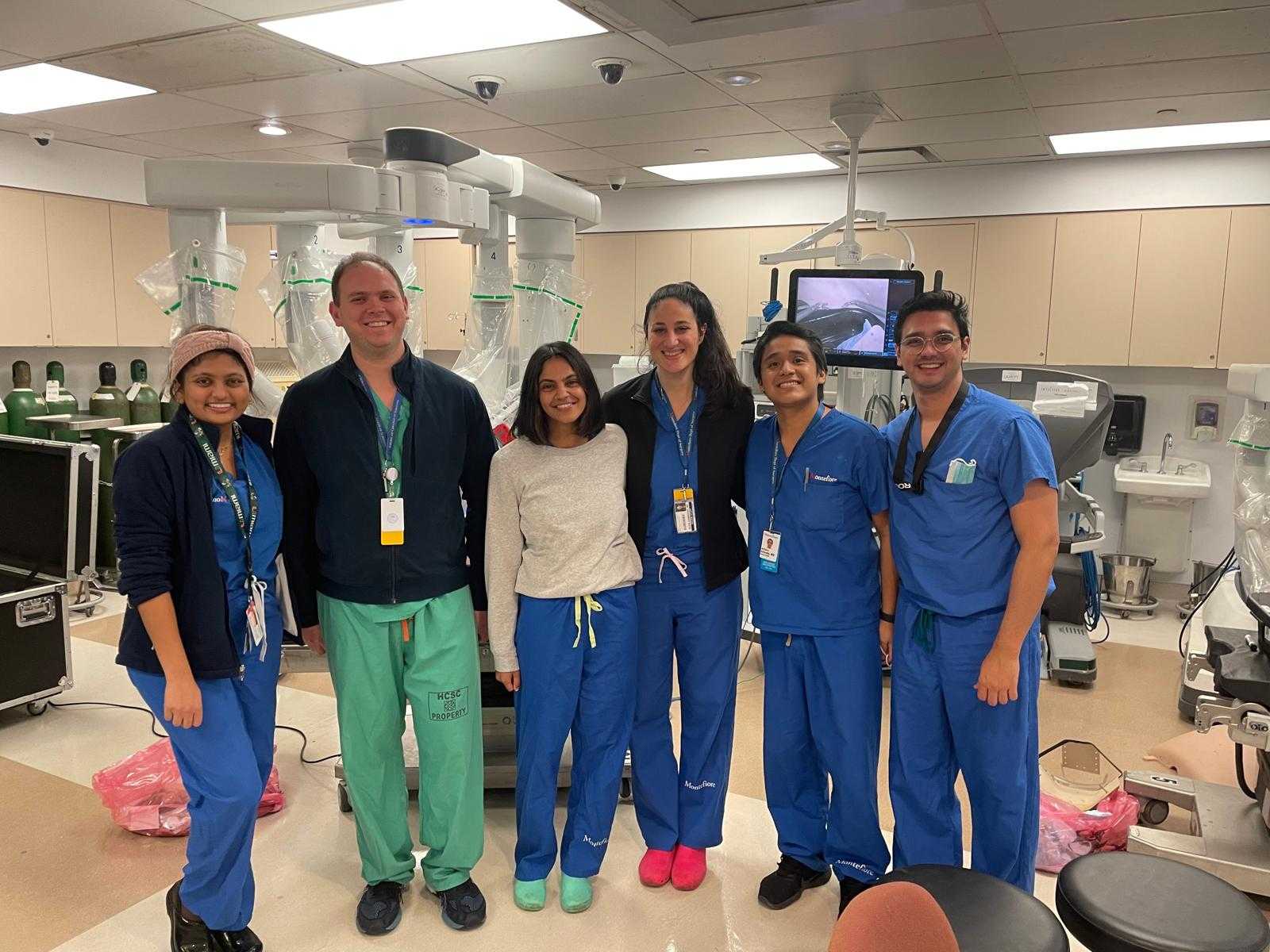 Group of medical professionals in scrubs and casual clothing standing together in a surgical training room with robotic equipment.