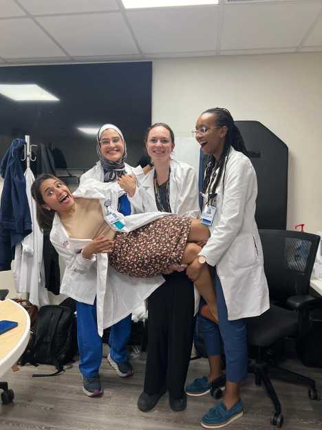 Three women in lab coats playfully holding up a smiling colleague in an office or clinical setting.