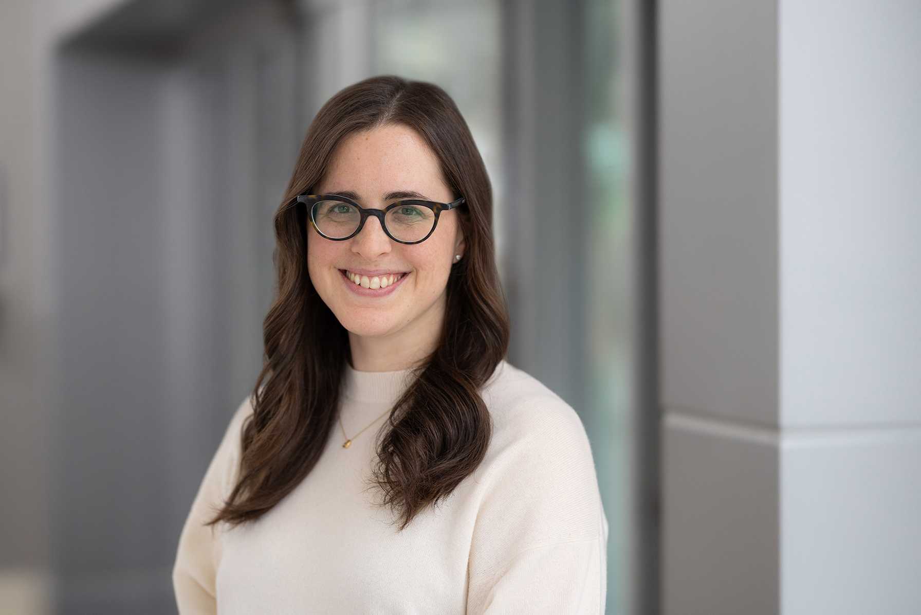 Portrait of a smiling woman with long brown hair and glasses, wearing a light sweater in a modern indoor setting.